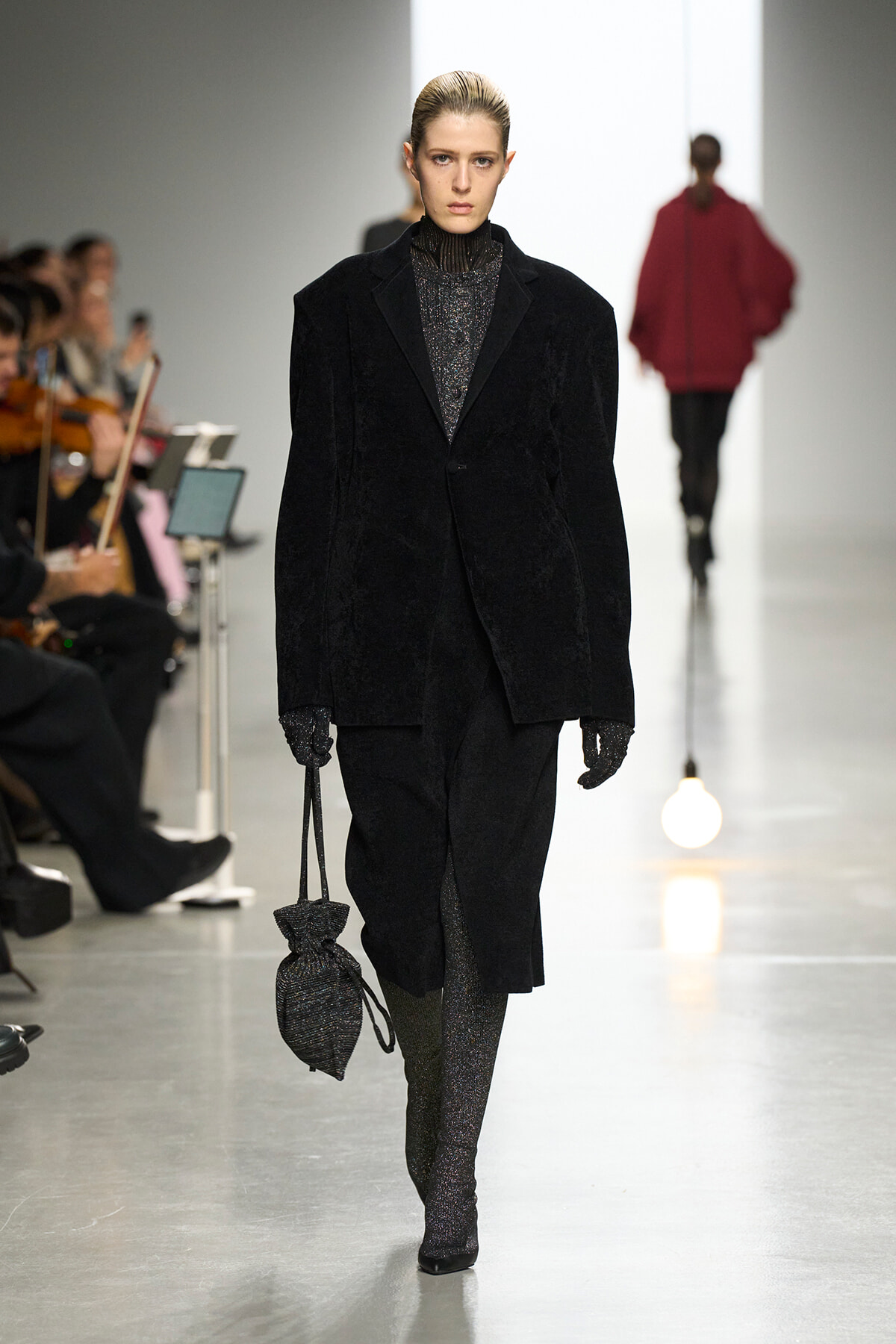 Model walking a fashion show in a black velvet coat over a glittery turtleneck, carrying a textured drawstring bag.