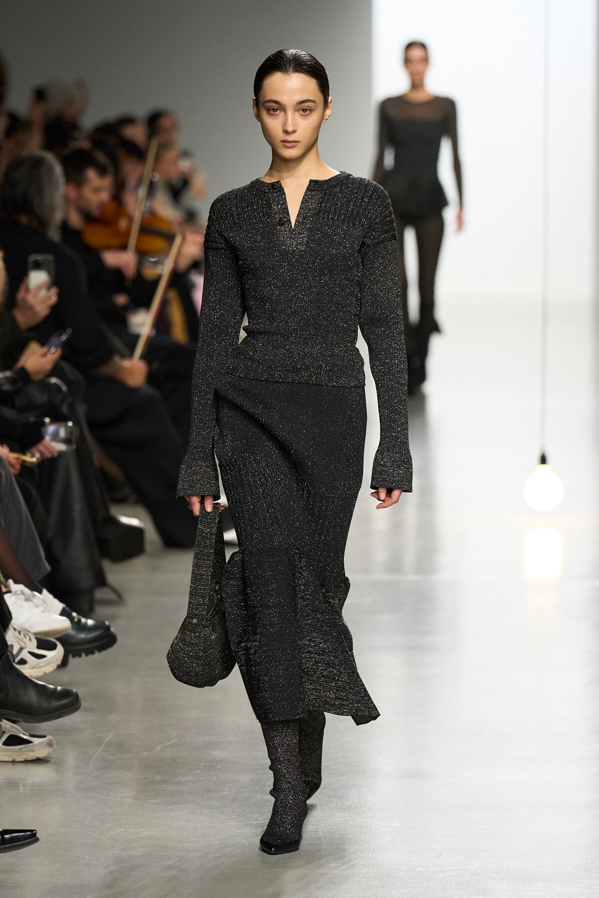 Model walking a glittery black knit outfit down the runway, carrying a small bag; audience seated along both sides of the catwalk.