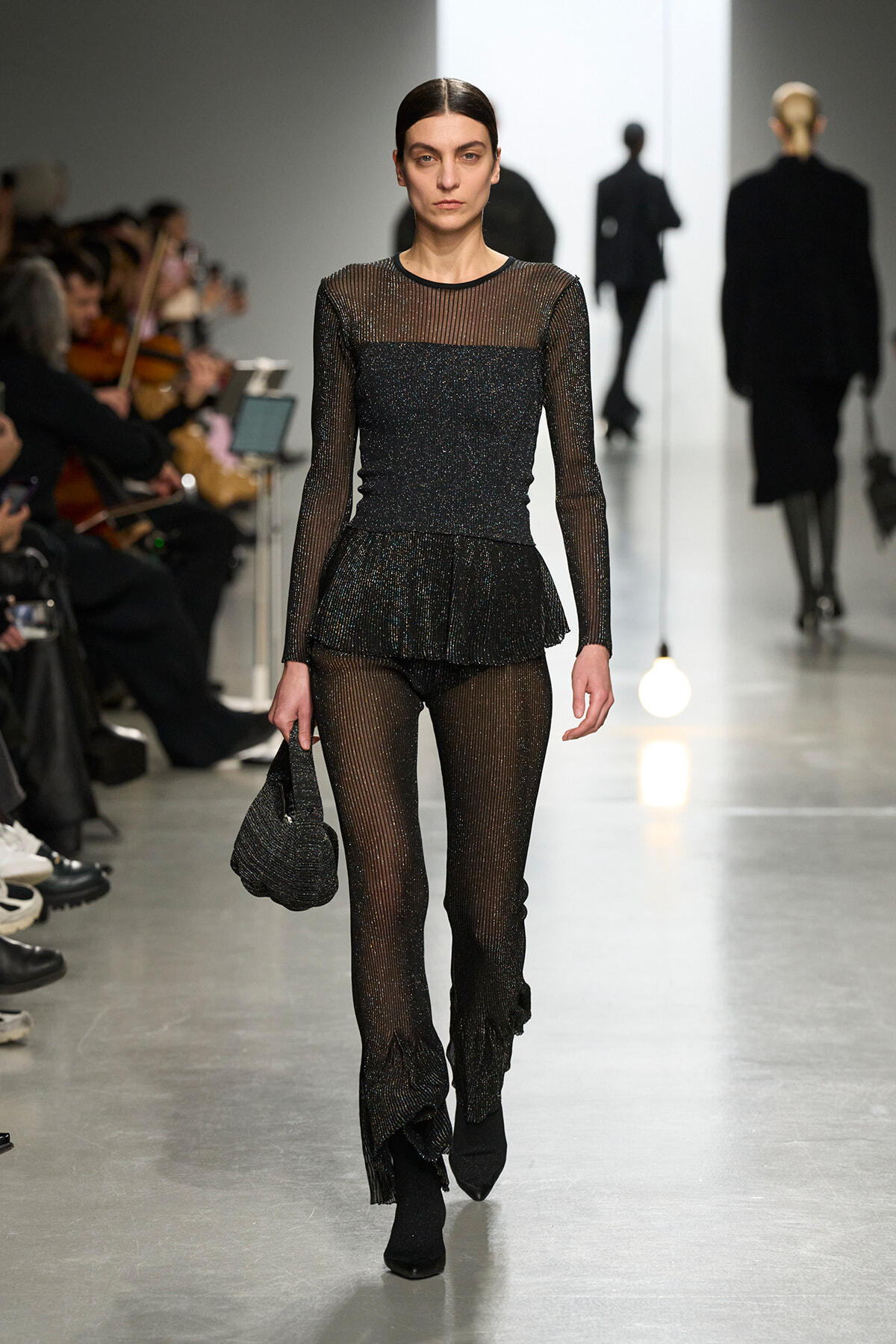 Model on a runway wearing a sheer black glittery top and matching pants, carrying a small black bag. g