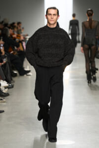 Male model walks down runway in a textured black sweater and black trousers at a fashion show.