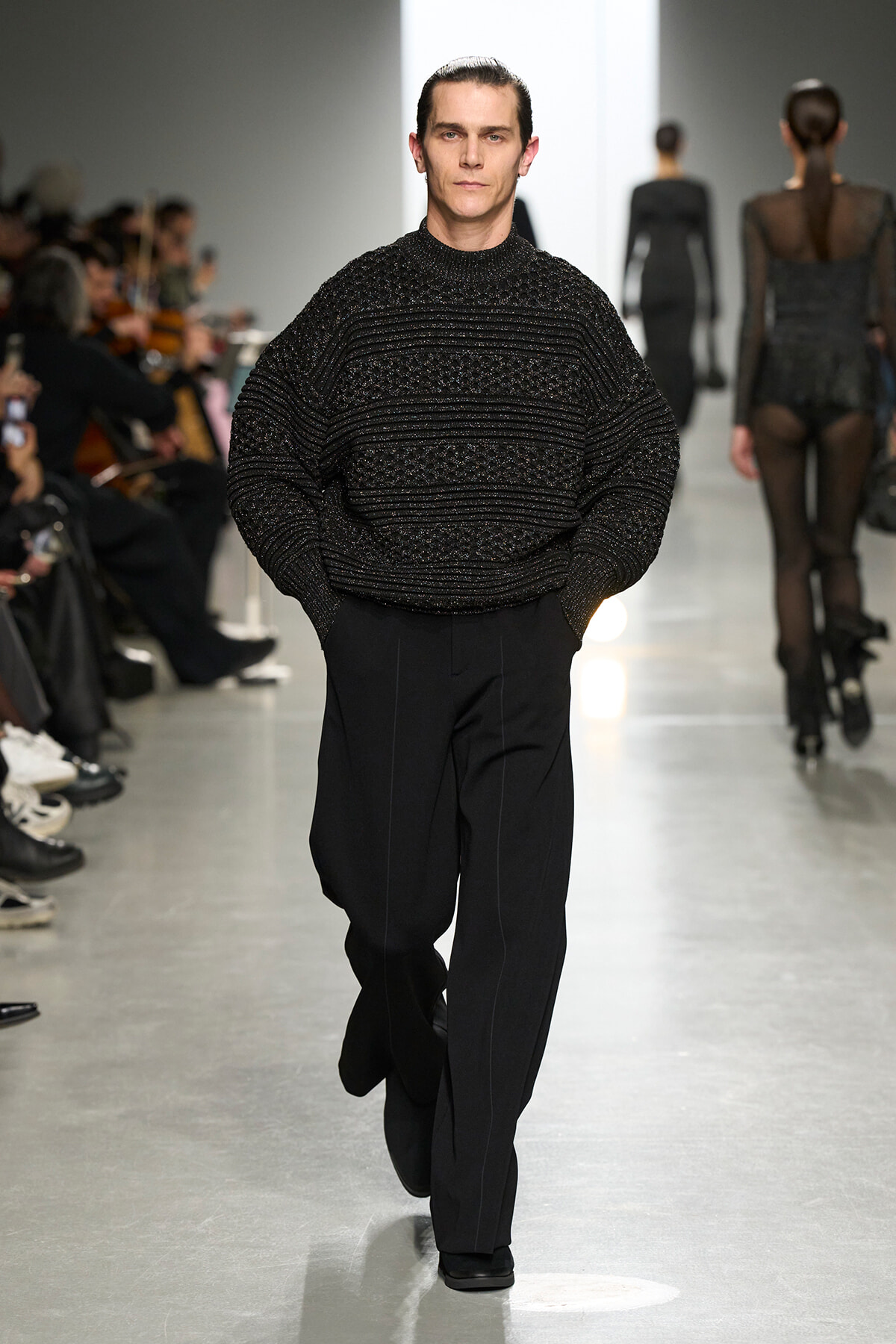 Male model walks down runway in a textured black sweater and black trousers at a fashion show.