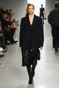 Model walking runway in a black blazer paired with a knee-length skirt covered in black circular sequins; audience visible on the sides.