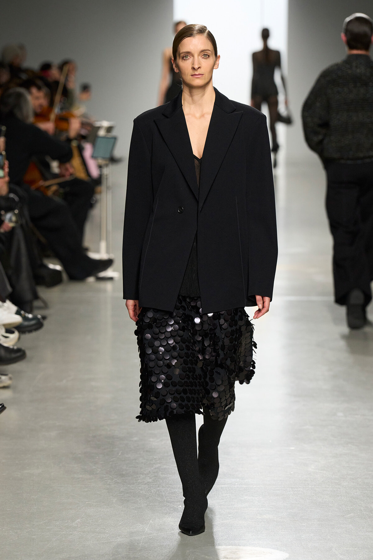 Model walking runway in a black blazer paired with a knee-length skirt covered in black circular sequins; audience visible on the sides.