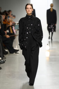 Model walks the runway wearing a black tailored suit with scattered round embellishments on the jacket and trousers, audience in the background