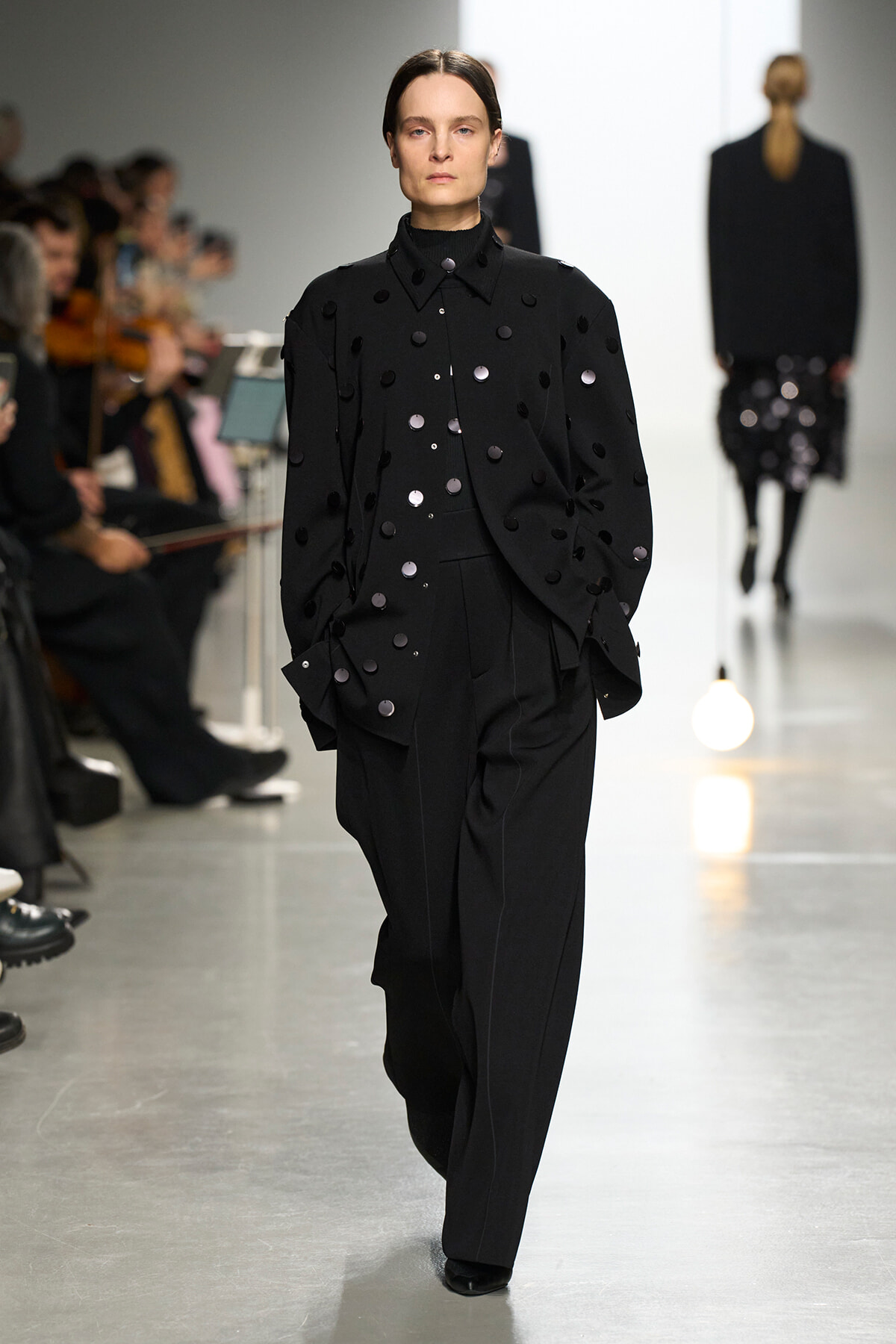 Model walks the runway wearing a black tailored suit with scattered round embellishments on the jacket and trousers, audience in the background