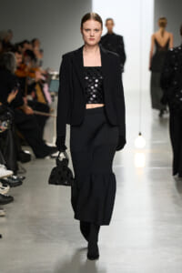 Model walks a fashion show in a black blazer over a sequined crop top, with a ribbed long skirt, gloves, and a small dotted handbag.
