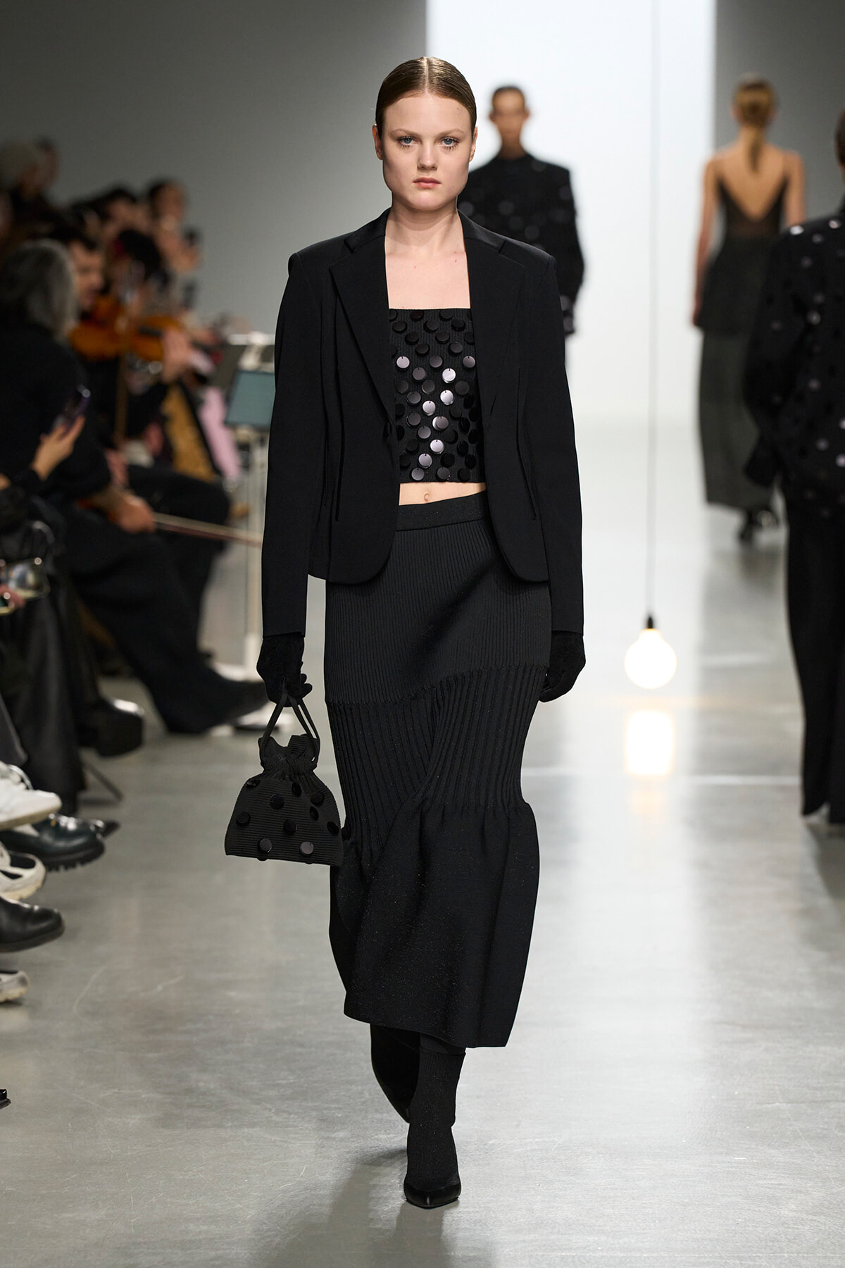 Model walks a fashion show in a black blazer over a sequined crop top, with a ribbed long skirt, gloves, and a small dotted handbag.