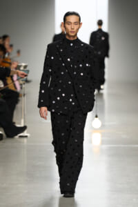 Model on a runway wearing a black suit covered in circular black and silver dots; audience blurred to the side.
