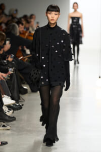 Model walks the runway in a black oversized coat covered with black and silver polka dots, wearing gloves and sheer black tights.