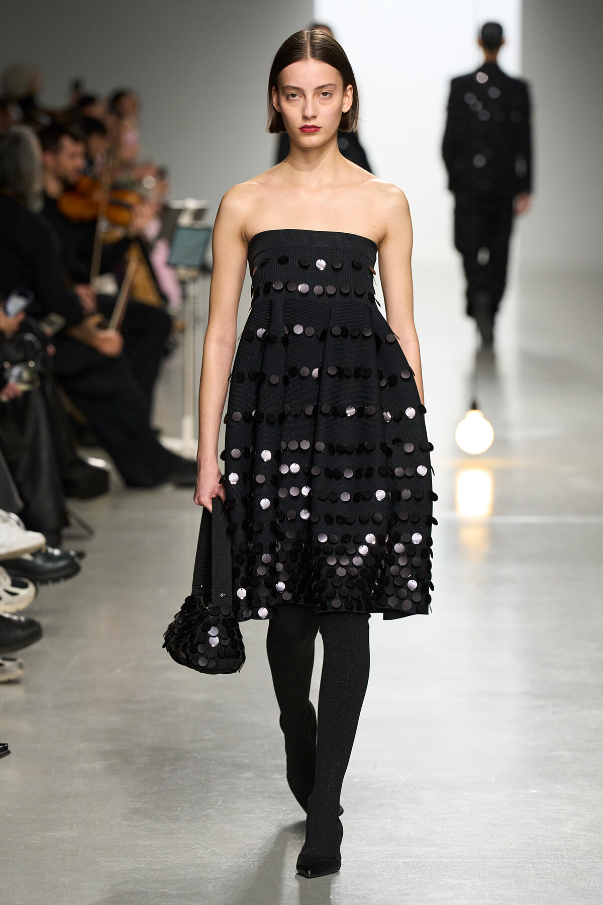 Model walks the runway in a strapless black dress covered in circular black sequins, carrying a matching bag.