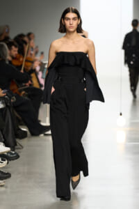 Model walks the runway in a black strapless top with a ruffled cape and wide-leg pants, audience visible to the side.