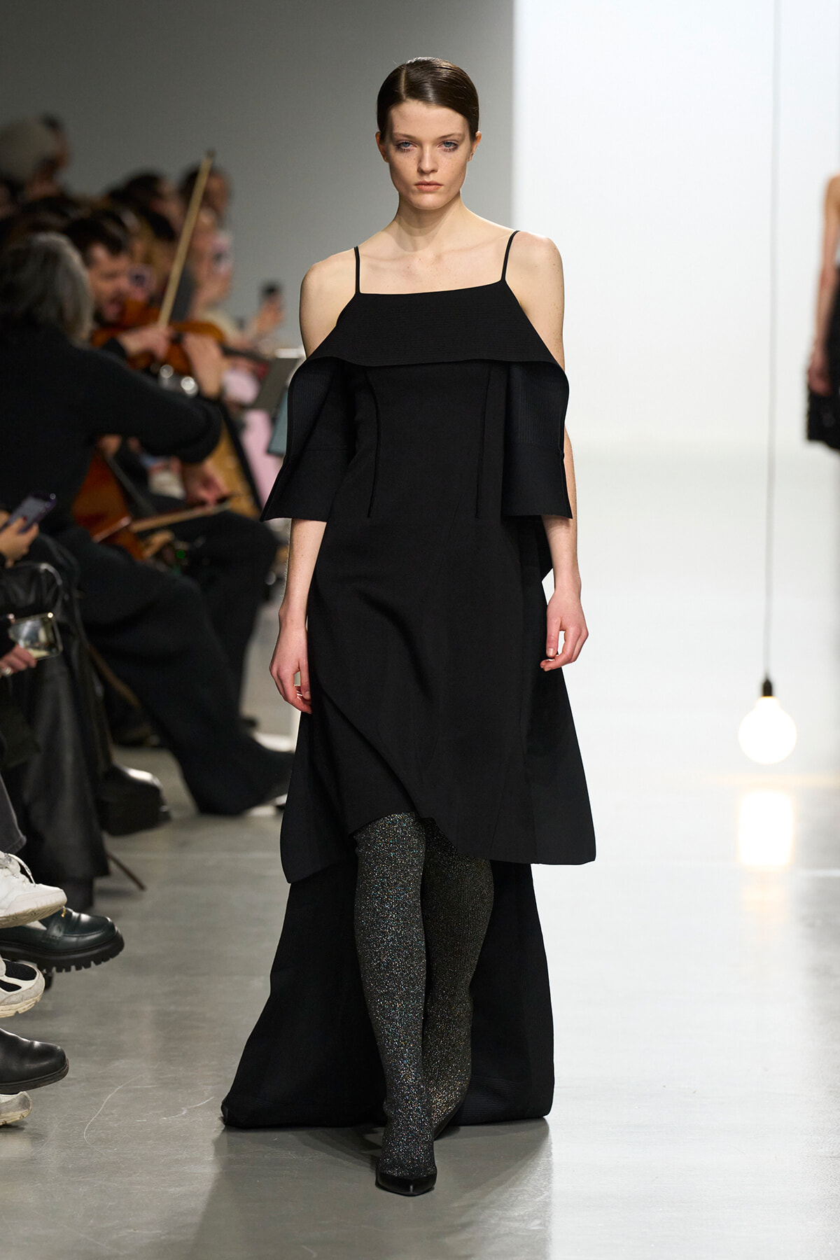 Model walks the runway in a black off-shoulder dress with a wide layered top and glittery tights, audience visible in the background.