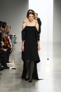 Model walks a fashion runway in a black off-the-shoulder dress with thin straps and glittery tights; audience sits along the left.