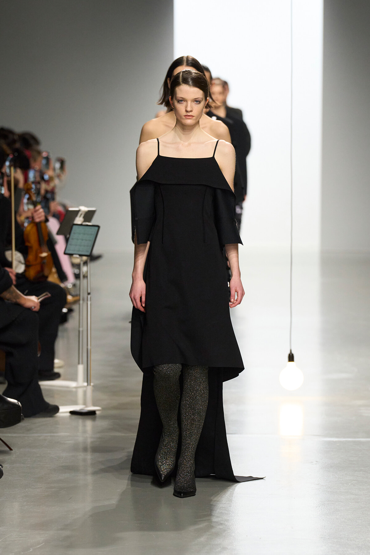 Model walks a fashion runway in a black off-the-shoulder dress with thin straps and glittery tights; audience sits along the left.