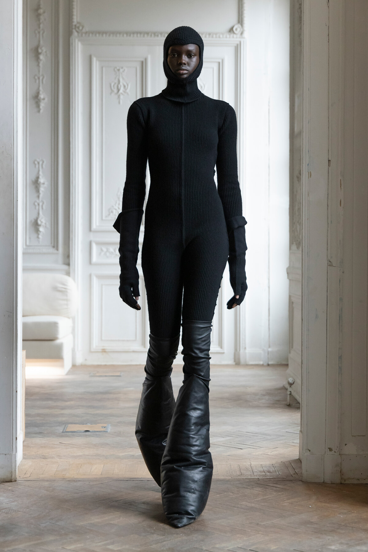 Person in a black full-body knit suit with hood and gloves stands in a white, ornate room.