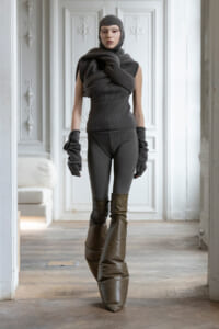 Person posing in a dark, layered knit outfit with a large scarf wrapped around the shoulders, knit cap, gloves, and tall brown boots in a bright room with ornate white doors behind them.