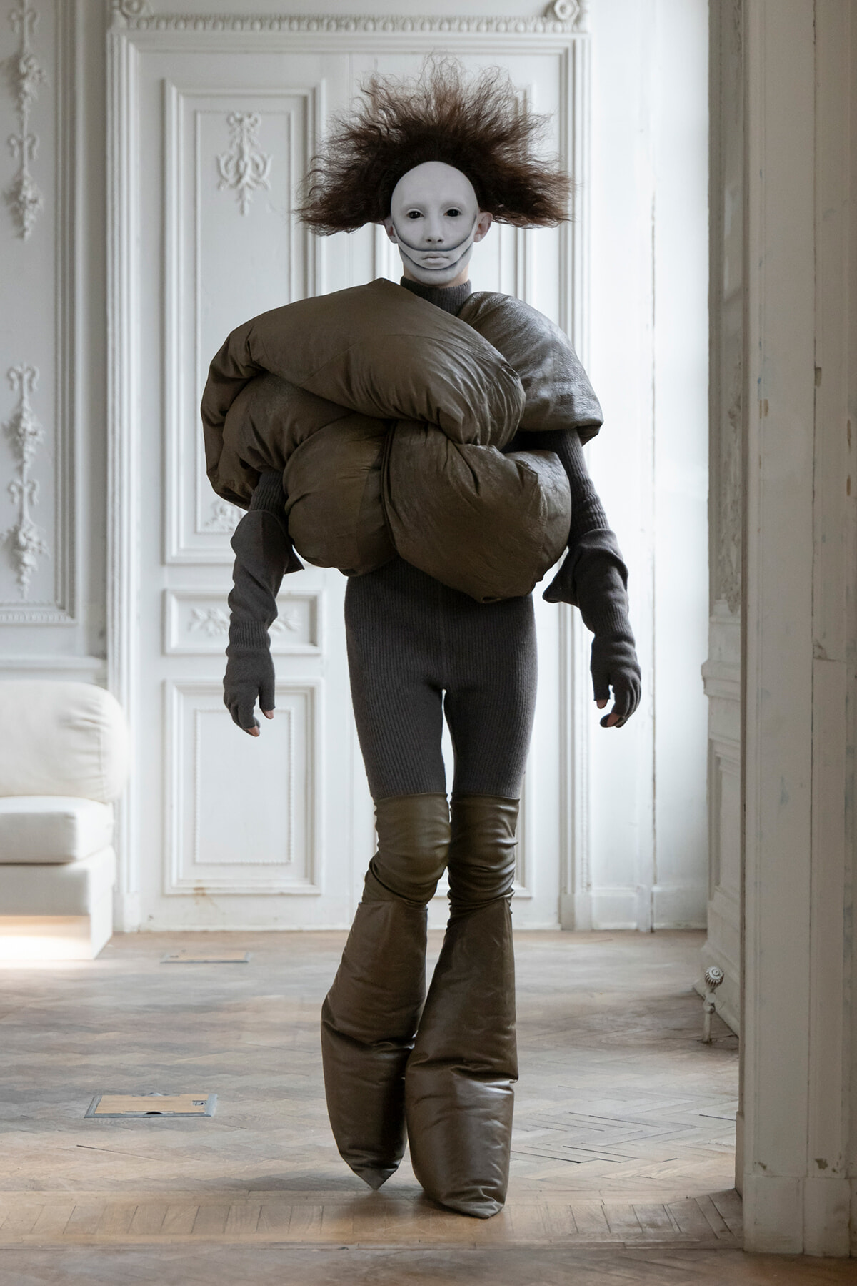 Person in a gray bodysuit with oversized brown inflatable padding and a white mask stands in a classical white room with ornate panels.