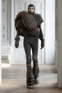 Person in a dark gray bodysuit with a large, brown, textured sculptural chest piece and tall, layered boots, standing in a light, ornate room.