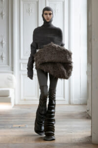 Full-body mannequin in a dark ribbed turtleneck and balaclava, carrying a voluminous brown fur-like skirt in a white ornate room.