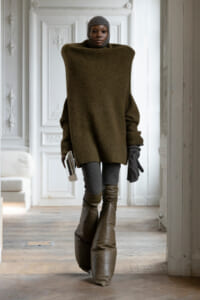 Person in an oversized olive sweater, knit cap, gloves, and tall boots, holding a small book in a decorative white room.