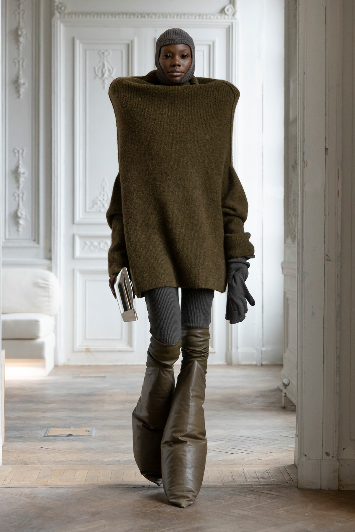Person in an oversized olive sweater, knit cap, gloves, and tall boots, holding a small book in a decorative white room.