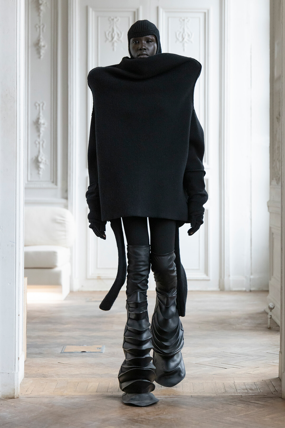 Person in an all-black ensemble with an oversized sweater, beanie, gloves, and tall sculpted boots stands in a white ornate interior.