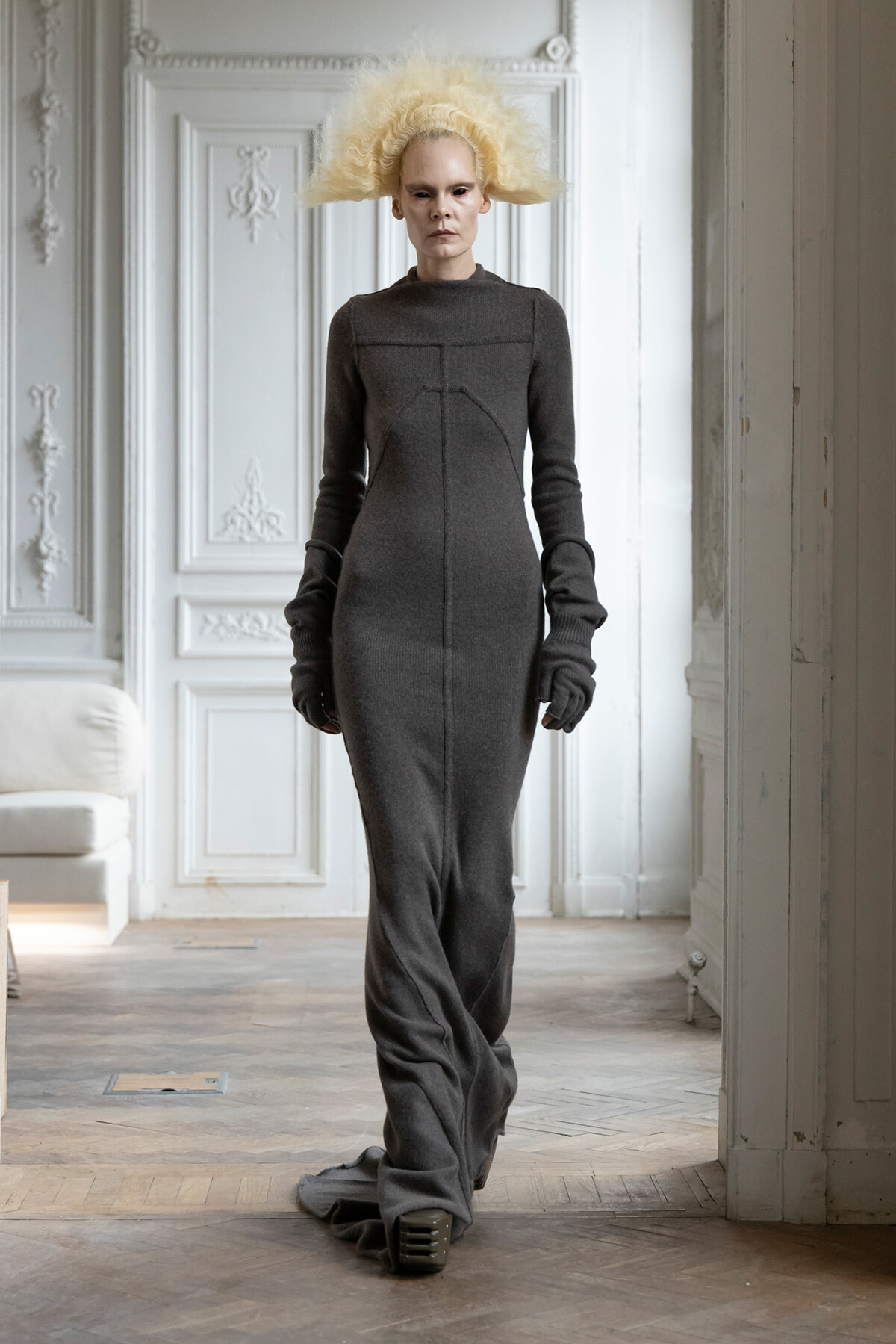 Model in a long dark grey knit dress with structured seams and a voluminous pale blonde wig, standing in an ornate white room.