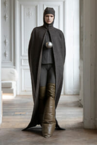 Model in a dark gray hooded cape with a large silver spherical pendant, indoors in a white ornate room.