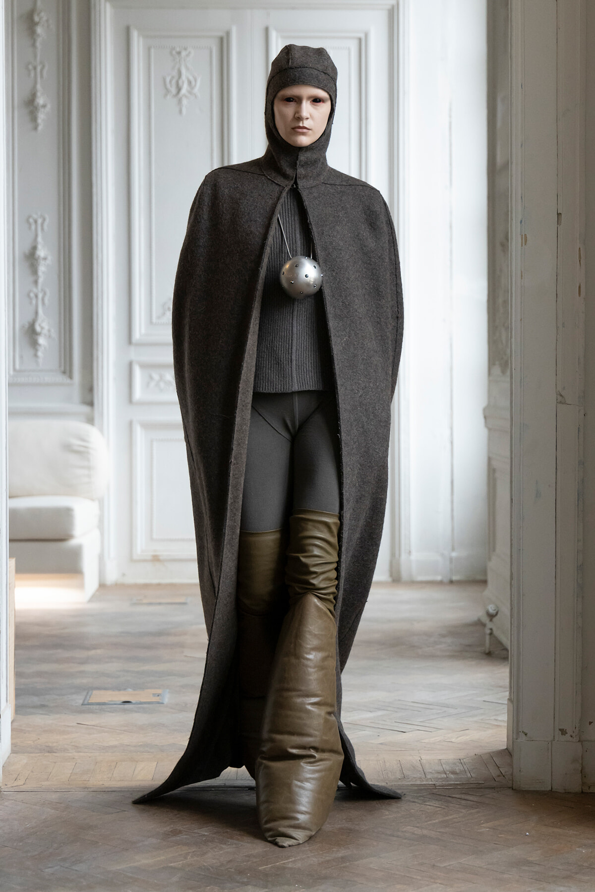 Model in a dark gray hooded cape with a large silver spherical pendant, indoors in a white ornate room.