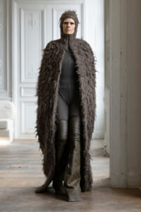 Person wearing a long shaggy brown fur coat with hood, black bodysuit, and tall boots, standing in a room with white ornate panels on the walls.