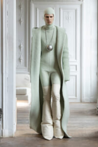 Model in a pale mint knit outfit: oversized coat and turtleneck with hood, white boots, and a large silver spherical pendant on a necklace, indoors with ornate white walls.