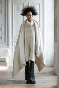 Model in an all-white ensemble: oversized cape over a ivory turtleneck sweater, paired with black tall boots in a white ornate room.