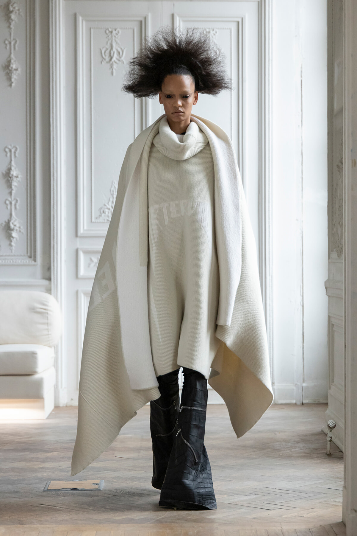 Model in an all-white ensemble: oversized cape over a ivory turtleneck sweater, paired with black tall boots in a white ornate room.