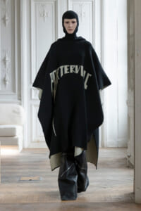 Model wearing a loose black poncho with a hooded knit cap, standing in a bright, ornate room with white paneled walls.