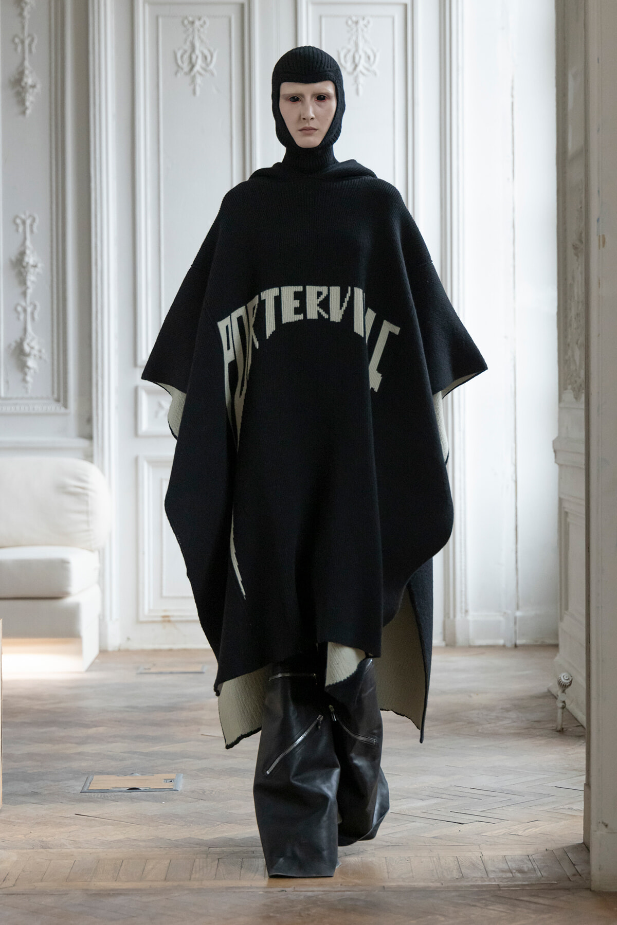 Model wearing a loose black poncho with a hooded knit cap, standing in a bright, ornate room with white paneled walls.