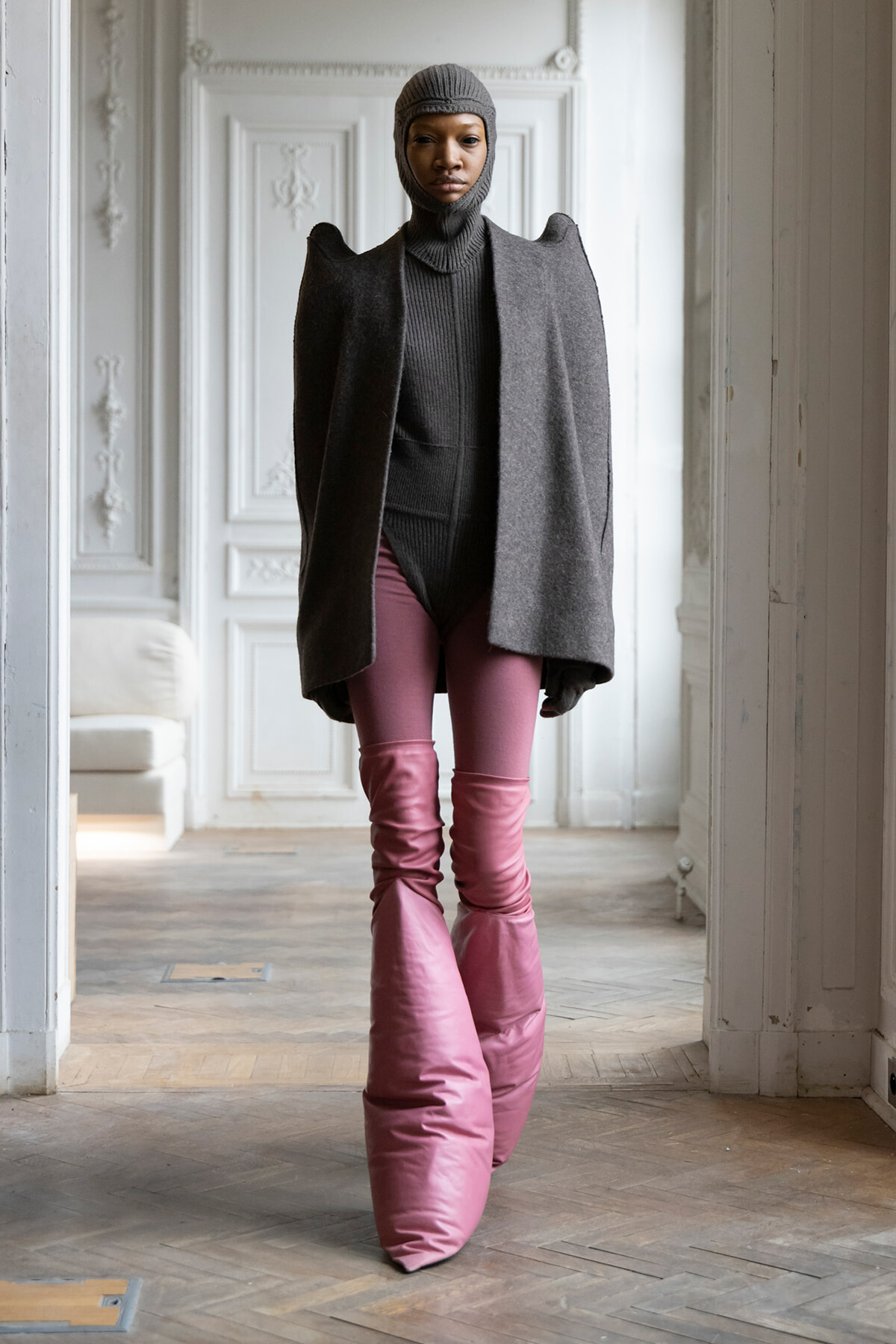 Model in a oversized gray cape coat over a ribbed turtleneck and balaclava, paired with bright pink thigh-high boots in a bright, ornate room.