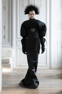 Model wearing an oversized black outfit with huge padded shoulders, posing in a white ornate room with paneled walls.
