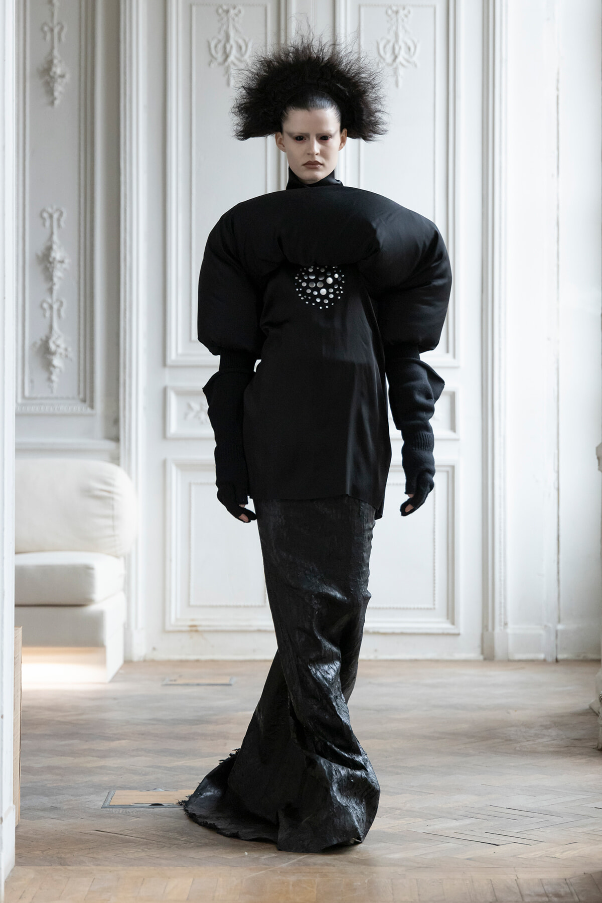 Model wearing an oversized black outfit with huge padded shoulders, posing in a white ornate room with paneled walls.