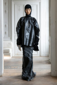 Full-body fashion shot of a person in a glossy black leather tunic, hooded headpiece, and gloves, standing in a bright, ornate room.