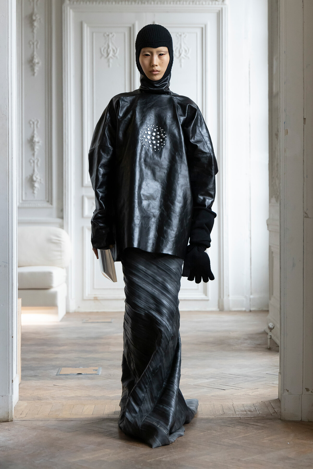 Full-body fashion shot of a person in a glossy black leather tunic, hooded headpiece, and gloves, standing in a bright, ornate room.