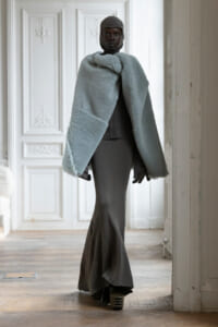 Model wearing a light gray faux-fur cape and knit balaclava in a pale, ornate room with paneled white doors behind her, full-length portrait.