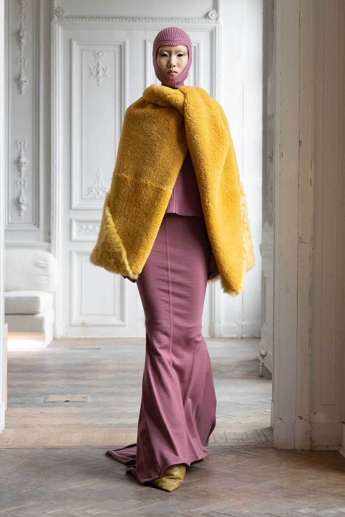 Model wearing a bright yellow faux-fur cape over a magenta knit outfit with a pink balaclava in a bright, ornate room.