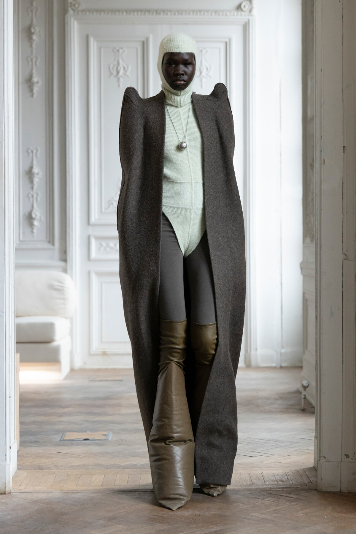 Model wearing a long dark cape over a mint green knit turtleneck and hat, standing in a white ornate room.
