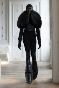 Full-body shot of a model wearing an oversized black puffed jacket with a zipper, standing in a white paneled room wearing black leggings and knee-high boots.