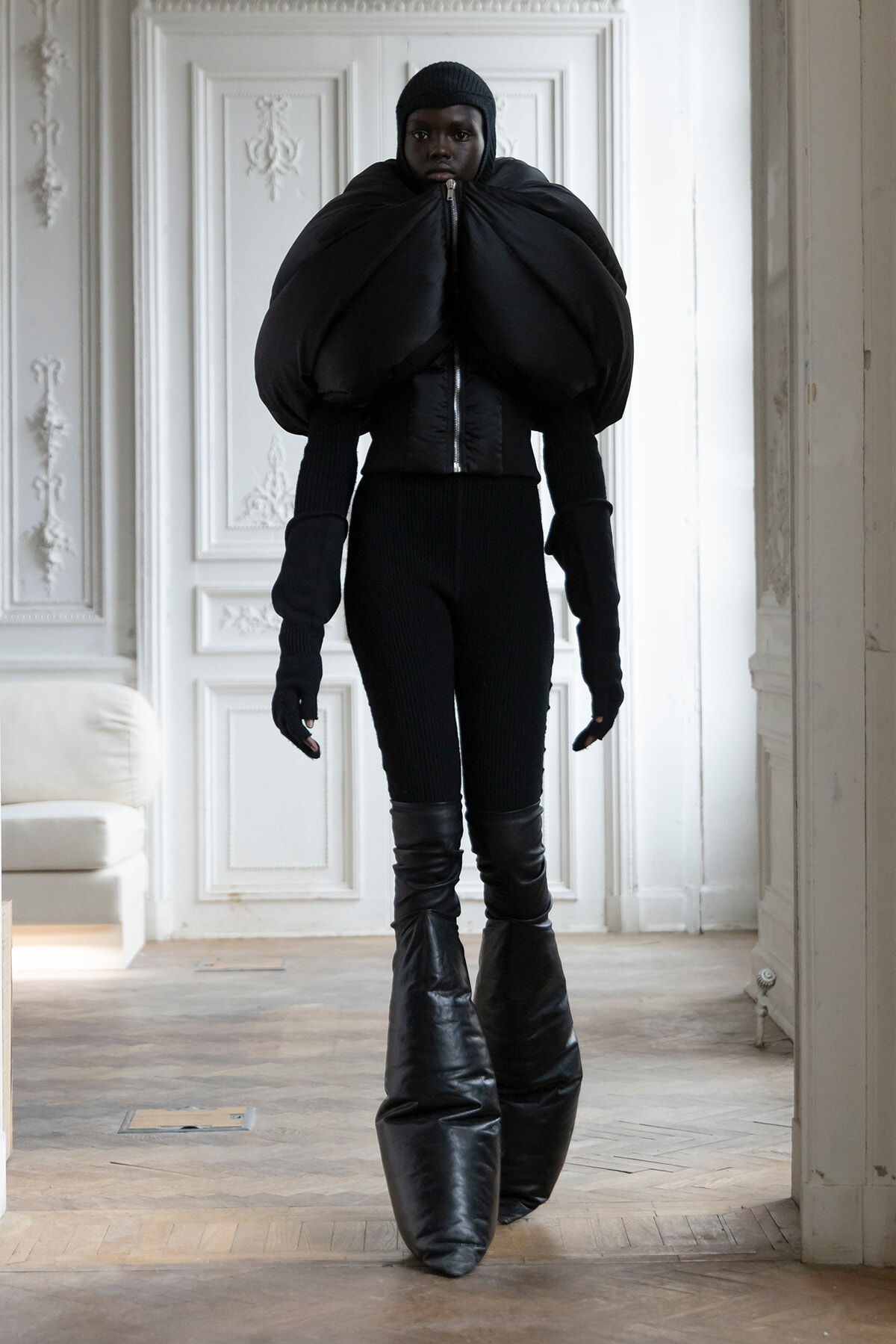 Full-body shot of a model wearing an oversized black puffed jacket with a zipper, standing in a white paneled room wearing black leggings and knee-high boots.