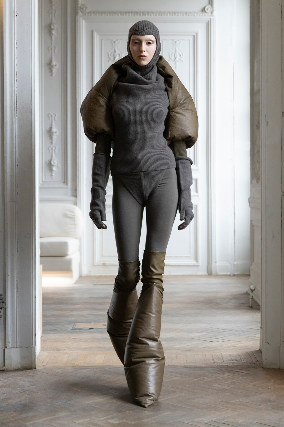 Person in a grey ribbed turtleneck bodysuit with a large brown puff shoulder cape, grey gloves, and brown knee-high boots standing in a pale ornate room.