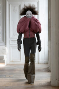 Person wearing a white mask and a dramatic burgundy padded garment with oversized shoulders, standing in a white ornate room.