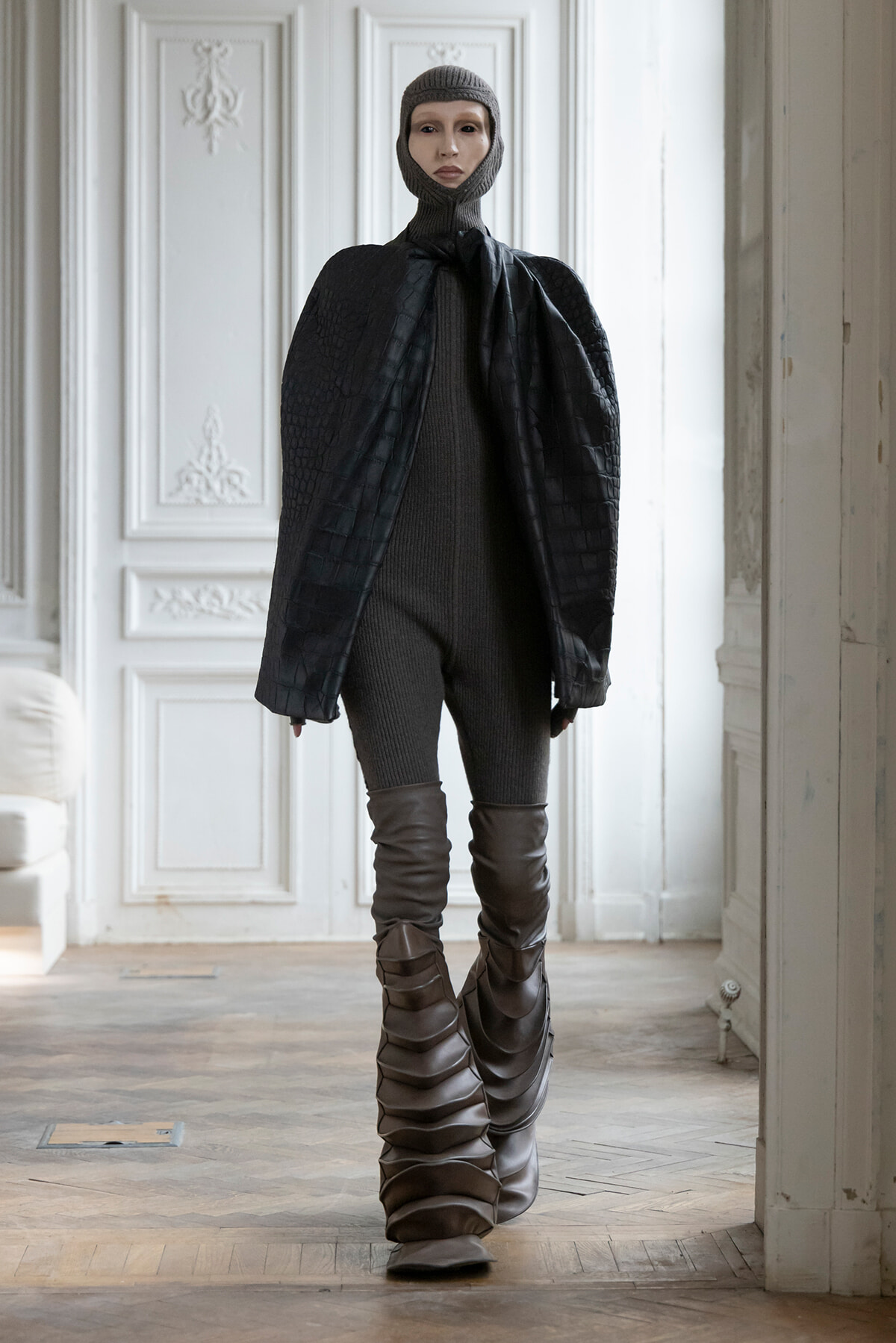 Model in a gray knit balaclava and oversized black quilted cape, standing in a bright ornate room with white panels, wearing a dark ribbed bodysuit and tall boots?