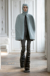 Model in a pale blue plush hooded cape, standing indoors with ornate white walls and tall sculptural boots.