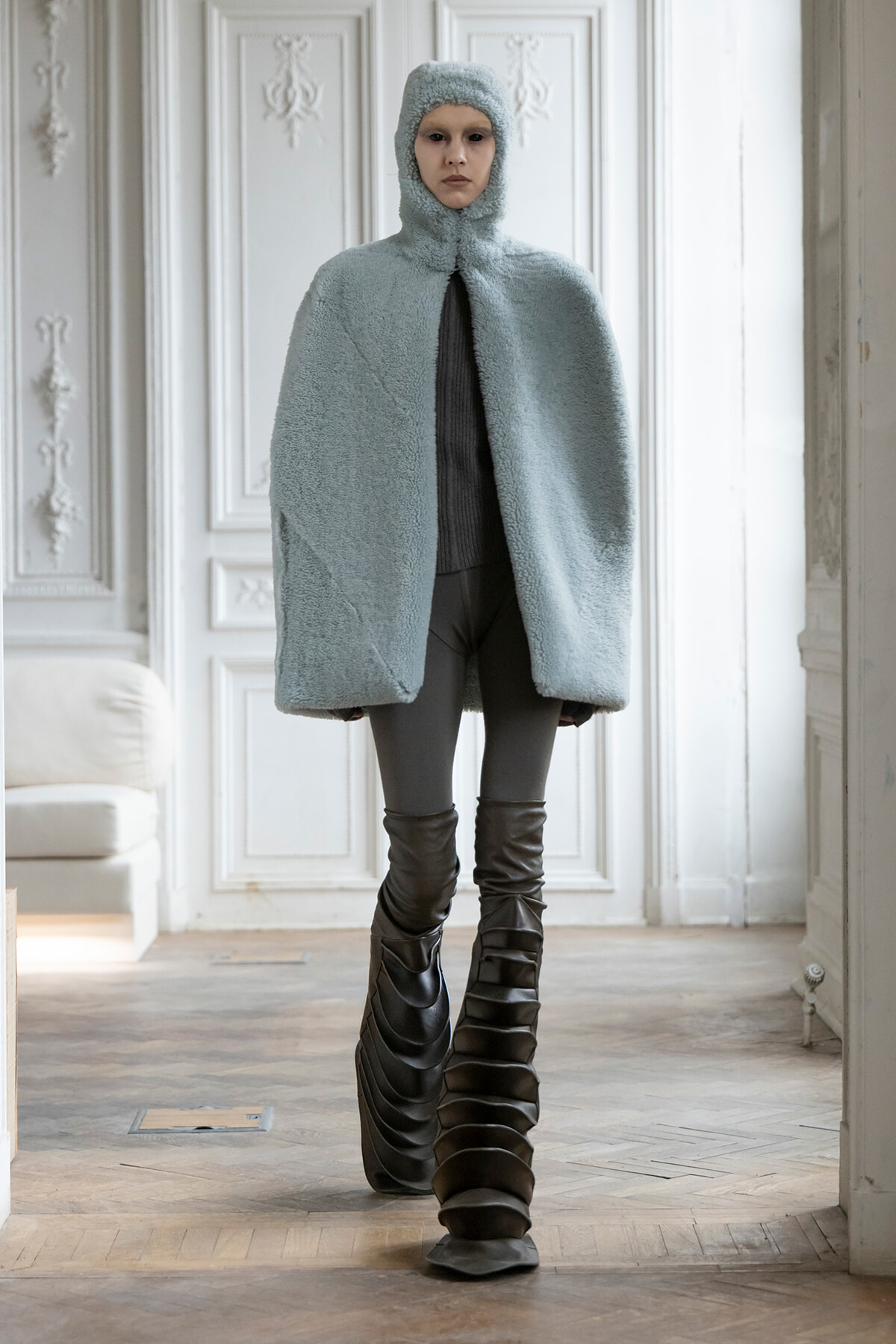 Model in a pale blue plush hooded cape, standing indoors with ornate white walls and tall sculptural boots.